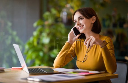 Photo for Attractive cheerful young businesswoman working at coffee shop, lady sitting at table alone, working on laptop, having phone conversation, holding eyeglasses, looking at copy space and smiling - Royalty Free Image