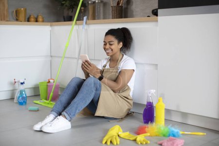 Téléchargez les photos : Glad millennial african american lady in apron sits on floor with cleaning supplies chatting on smartphone, sits on floor in kitchen interior. Blog, rest from household chores with device at home - en image libre de droit