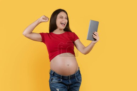Foto de Excited Young Pregnant Woman Holding Digital Tablet And Celebrating Success, Cheerful Expectant Female Raising Fist And Exclaiming With Joy While Standing Isolated Over Yellow Background, Copy Space - Imagen libre de derechos