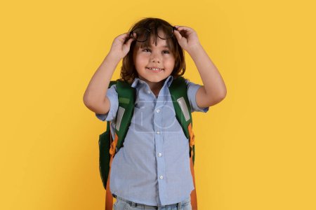 Foto de Kids and eyesight problem. Cute little school boy with backpack taking off his eyeglasses on forehead, having some troubles with vision, orange studio background - Imagen libre de derechos