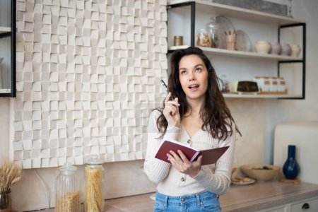Foto de Excited lady taking notes, holding notepad and pen, writing new recipes to her cooking book, standing in kitchen interior, copy space. Cheerful woman planning her day, writing tasks - Imagen libre de derechos