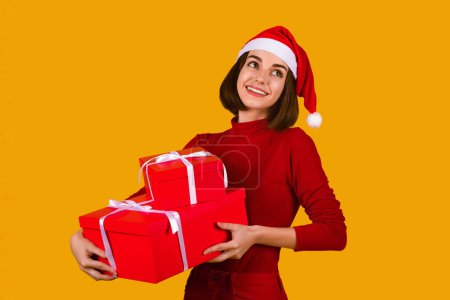 Foto de Happy beautiful young woman in Santa hat and red outfit holding xmas presents and looking at copy space, advertisement for Christmas sales or discounts, orange studio background - Imagen libre de derechos