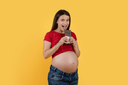Téléchargez les photos : Portrait Of Cheerful Young Pregnant Lady Biting Chocolate Bar And Looking At Camera While Posing Isolated Over Yellow Background, Happy Expectant Woman Enjoying Eating Sweets During Pregnancy - en image libre de droit