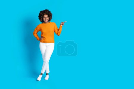 Téléchargez les photos : Full length studio shot of stylish black millennial lady in nice outfit posing on blue background, pointing at copy space and smiling, african american woman showing nice offer - en image libre de droit
