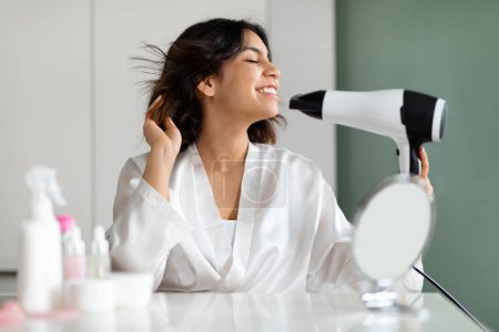 Photo for Cheery happy emotional brunette young middle eastern woman having fun while styling hair at home, female in silk bathrobe sitting at vanity table in front of mirror, using hairdryer - Royalty Free Image