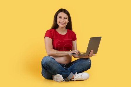 Foto de Remote Work During Pregnancy. Happy Pregant Female Sitting On Floor With Laptop, Smiling Expectant Lady Using Computer For Online Job While Posing Over Yellow Studio Background, Copy Space - Imagen libre de derechos