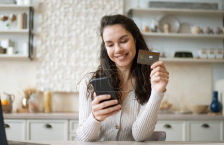 Photo for Grocery delivery application. Happy woman holding credit card and using cellphone, ordering food and products, sitting in kitchen. Online shopping app - Royalty Free Image