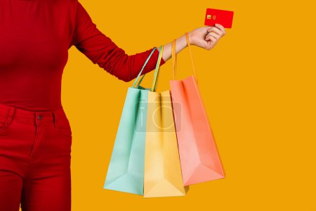 Photo for Unrecognizable woman in red outfit holding colorful shopping bags and red plastic credit card over orange studio background, copy space, cropped shot. Unlimited shopping, retail concept - Royalty Free Image