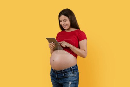 Foto de Smiling Pregnant Woman Using Digital Tablet While Standing Over Yellow Background In Studio, Happy Young Expectant Female Reading Pregnancy Blog Online Or Shopping In Internet, Copy Space - Imagen libre de derechos