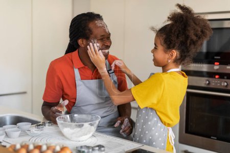 Téléchargez les photos : Cheerful Black Girl Playing With Dad In Kitchen While They Baking Muffins Together, Happy African American Family Father And Daughter Having Fun At Home, Child Powdering Daddys Face With Flour - en image libre de droit