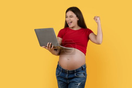 Foto de Joyful Young Pregnant Woman Holding Laptop Computer And Celebrating Success, Cheerful Expectant Female Raising Fist And Exclaiming With Excitement While Standing Over Yellow Background, Copy Space - Imagen libre de derechos