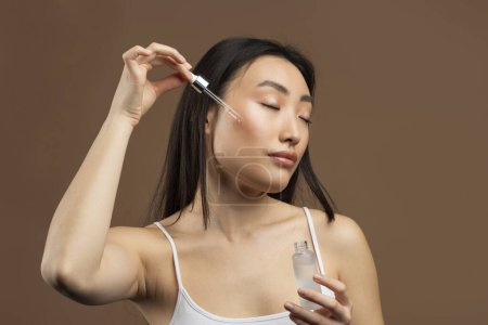 Téléchargez les photos : Beautiful asian woman applying face serum with dropper while posing over brown studio background. Young asian lady moisturizing skin, enjoying new beauty product - en image libre de droit