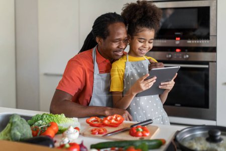 Téléchargez les photos : Happy Black Girl And Her Dad Using Digital Tablet In Kitchen, African American Father And Cute Preteen Female Child Searching Recipe Online While Cooking Lunch Together At Home, Free Space - en image libre de droit