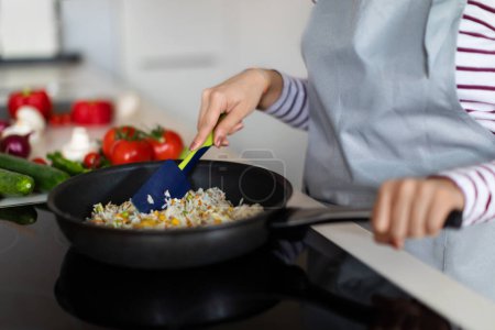 Téléchargez les photos : Closeup shot of female hands holding cooking spatula, mixing rice with vegetables in frying pan on electric stove, cropped of woman preparing food at kitchen, copy space. Healthy diet, nutrition - en image libre de droit