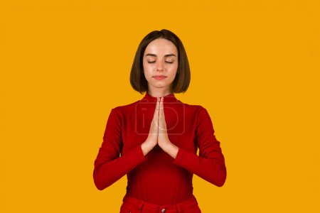 Téléchargez les photos : Calm spiritual brunette young woman praying with closed eyes. Serious peaceful lady with joining hands meditating. Isolated on orange studio background. Belief concept, copy space - en image libre de droit