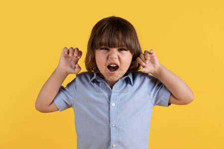 Téléchargez les photos : Kids aggression. Close up portrait of angry little boy threatening to camera, showing fingers like claws and frowning face, orange studio background - en image libre de droit