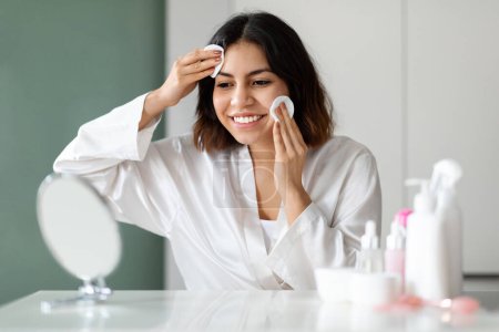 Téléchargez les photos : Face care routine. Smiling beautiful young middle eastern woman in white bathrobe looking at mirror and using cotton pads for makeup removal or face cleansing, home interior, copy space - en image libre de droit