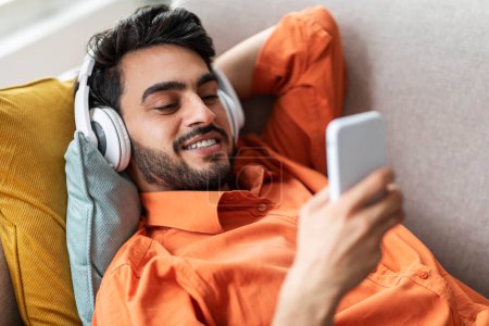 Photo for Candid shot of relaxed attractive bearded middle eastern millennial man lying on sofa at home, using phone and newest wireless headphones, waching video on Internet, using mobile app, closeup - Royalty Free Image