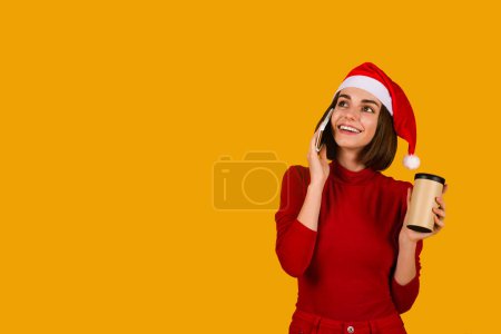 Photo for Chreerful pretty young brunette woman in red outfit and Santa hat drinking coffee to go, having phone conversation, looking at copy space and smiling, orange studio background - Royalty Free Image