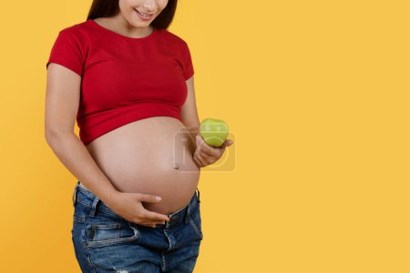 Foto de Pregnancy Diet. Young pregnant female holding green apple and embracing her belly, smiling expectant woman enjoying healthy nutrition, standing isolated over yellow background, copy space - Imagen libre de derechos