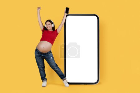 Foto de Great App. Joyful Pregnant Woman On Headphones Standing Near Big Blank Smartphone With White Screen Over Yellow Background, Happy Expectant Female Recommending Music Streaming Application, Mockup - Imagen libre de derechos