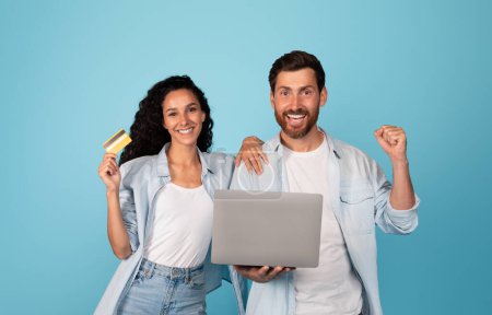 Téléchargez les photos : Smiling young european couple with credit card and laptop make gesture of victory and success, great purchase, sale, isolated on blue background, studio. Online shopping for shopaholics and cashback - en image libre de droit