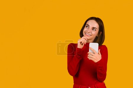 Foto de Excited pretty young brunette woman in red checking newest mobile application, using modern cell phone and looking at copy space over orange studio background, touching her chin and smiling - Imagen libre de derechos
