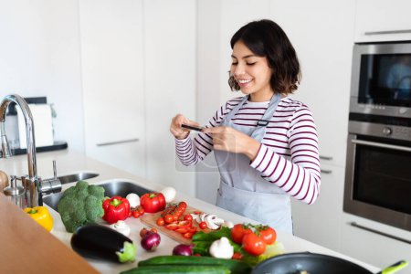 Photo for Food blogging concept. Smiling pretty hispanic young woman taking photo of healthy vegetables on cellphone, cooking fresh salad while standing in kitchen. Nutrition and weight loss blog concept - Royalty Free Image