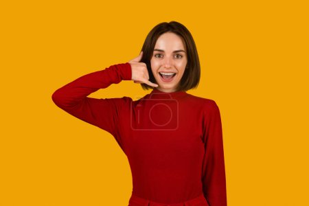 Téléchargez les photos : So call me maybe. Friendly cheerful attractive young woman in red showing a call me sign and smiling while standing isolated on orange background, studio shot, copy space for advertisement - en image libre de droit