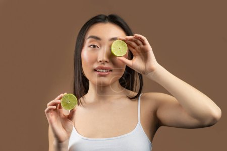 Téléchargez les photos : Fruit acids for skin care. Beautiful asian lady holding lime halves in front of her face on brown studio background. Young korean woman pampering herself with natural organic products - en image libre de droit