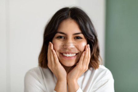 Téléchargez les photos : Headshot of happy smiling attractive young arabic lady in white homewear touching her cheeks, enjoying silky skin after using luxury organic premium beauty face care products, closeup - en image libre de droit