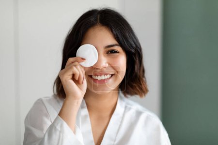 Foto de Closeup photo of cheerful happy young arabic woman wearing white silk bathrobe cover one eye with cotton pad and smiling, home interior, copy space. Face cleansing, skin care routine concept - Imagen libre de derechos