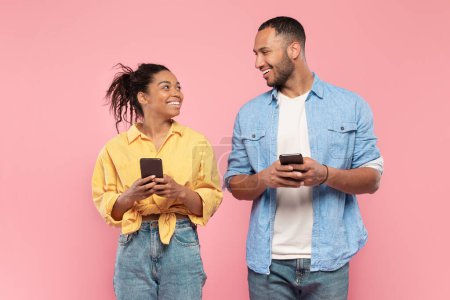 Photo for Cheerful black couple holding smartphones and looking at each other, excited man and woman using cellphones while standing together over pink background - Royalty Free Image