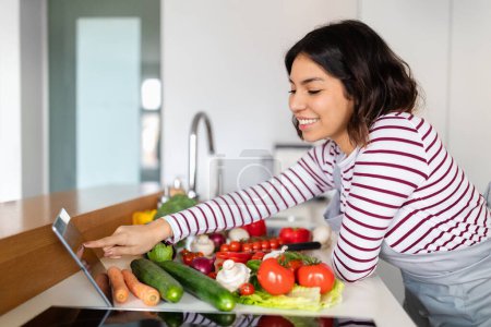 Photo for Vegan food blog, vlog concept. Cheerful smiling brunette young woman in casual outfit enjoy cooking at home, choosing nice recipe on Internet, using digital tablet, kitchen interior, copy space - Royalty Free Image