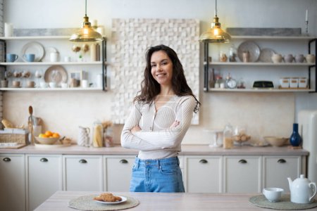 Foto de Portrait of happy young caucasian housewife posing with folded arms in kitchen interior, smiling and looking at camera while spending time at home alone, free space - Imagen libre de derechos