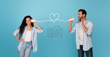 Photo for Smiling young european male, arab lady talking through wire in shape of heart, have fun and call, isolated on blue background, studio, panorama. Love, relationship, romance and distance communication - Royalty Free Image