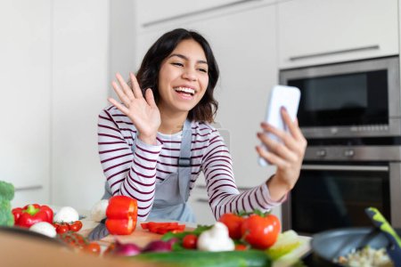 Foto de Cool friendly cheerful pretty brunette young woman influencer wearing apron streaming while cooking healthy tasty food at cozy modern kitchen, waving at smartphone screen, copy space - Imagen libre de derechos