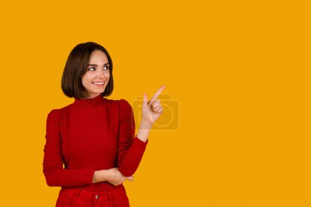 Foto de Cheerful pretty young caucasian woman with cute hairstyle in red pointing at copy space for advertisement and smiling over orange studio background, web-banner. Nice offer, discount, news - Imagen libre de derechos