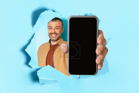Téléchargez les photos : Happy middle aged man holding big smartphone with blank screen in hand, showing gadget with copy space for mockup through torn blue paper hole, collage - en image libre de droit