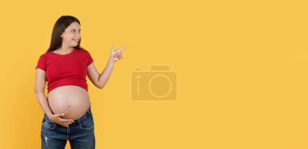 Foto de Nice Offer. Smiling Young Pregnant Lady Pointing Aside At Copy Space Over Yellow Background, Happy Beautiful Expectant Woman Demonstrating Empty Place For Advertisement And Touching Belly, Panorama - Imagen libre de derechos