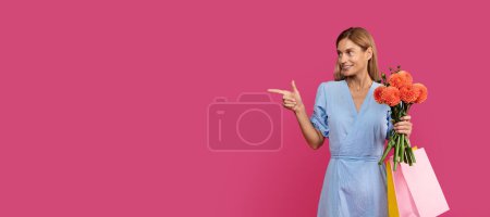 Téléchargez les photos : Happy adult caucasian woman blonde in dress with lot of packages with purchases and bouquet of flowers points finger at empty space, isolated on pink background, studio. Ad and offer, sale, shopping - en image libre de droit