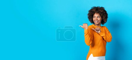 Téléchargez les photos : Cool smiling attractive african american young woman in casual with cute bushy hair aiming at copy space with thumbs on blue studio background, pointing at advertisement, web-banner - en image libre de droit