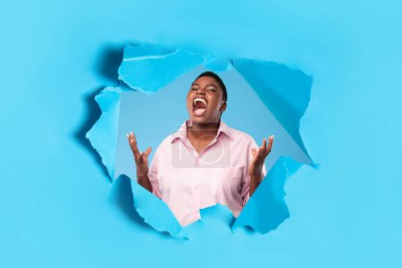 Photo for Angry oversize black woman shouting freaking out posing in hole of torn paper on blue studio background. Negative emotion, madness and anger concept - Royalty Free Image