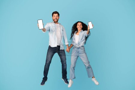Foto de Cheerful excited surprised millennial diverse guy and lady in casual with phones with blank screens jump, freeze in air, isolated on blue background. Fun together, offer and ad, great app and device - Imagen libre de derechos