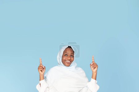 Foto de Check this. Happy black woman in hijab pointing up at copy space above her head, muslim female demonstrating free place for your ad on blue studio background, crop - Imagen libre de derechos