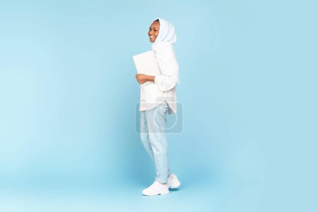 Foto de Full length shot of black muslim woman holding laptop walking over blue studio background, free copy space. Internet business and freelance career concept - Imagen libre de derechos