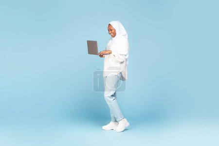 Foto de Positive african american muslim woman in hijab posing with laptop over blue studio background, full length shot. Islamic lady using computer for remote work or online shopping, free space - Imagen libre de derechos