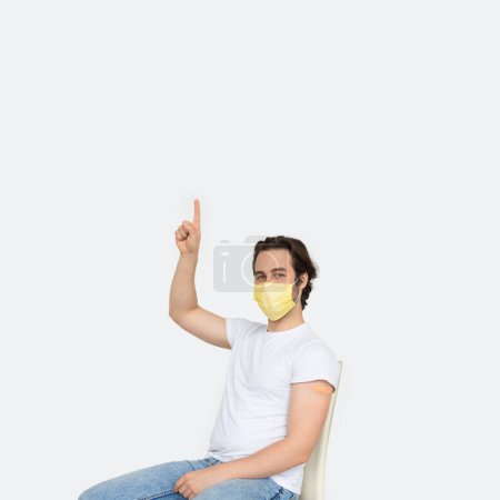 Photo for Young caucasian male in protective mask with band-aid on his shoulder after vaccination shows finger up on empty space, isolated on white background. Recommendation and advice, health care, medicine - Royalty Free Image