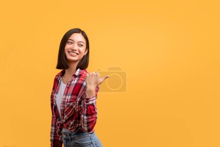 Photo for Check this. Happy asian lady looking and pointing back at copy space, positive korean woman indicating empty space for your ad, posing over yellow studio background - Royalty Free Image