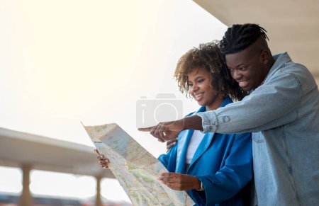 Foto de Smiling black couple looking at map while standing at railway station, happy young african american spouses checking new travel destination while waiting for train on platform, closeup shot - Imagen libre de derechos